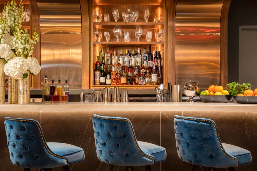 Tufted velvet stools at a long bar with backlit spirits bottles and moody lighting