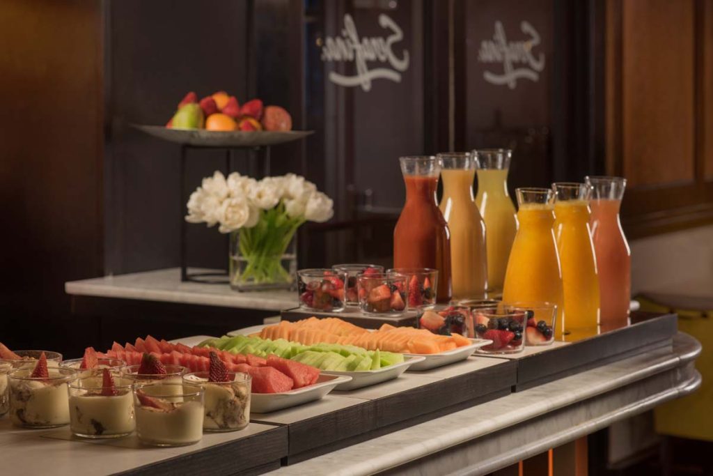 A buffet spread featuring several kinds of juice in glass carafes and different types of fruit on white platters